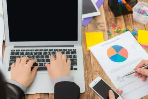 Como avaliar o desempenho de uma campanha publicitária digital: mãos digitando em notebook ao lado de gráficos, relatórios e celular em mesa de trabalho