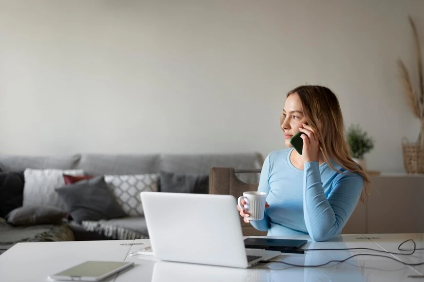 Mulher em home office falando ao telefone, com laptop e xícara na mesa, representa a flexibilidade do trabalho remoto