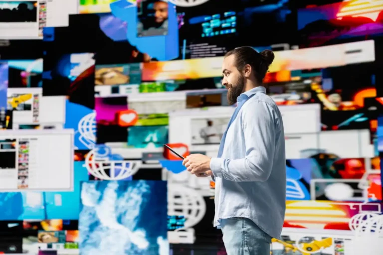Homem com barba e cabelo preso em um coque, usando camisa clara, segurando um tablet enquanto observa várias telas e gráficos de publicidade digital.