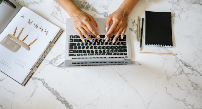 Vista superior de mãos digitando em laptop sobre mesa de mármore com caderno, lápis e revista aberta ao lado.