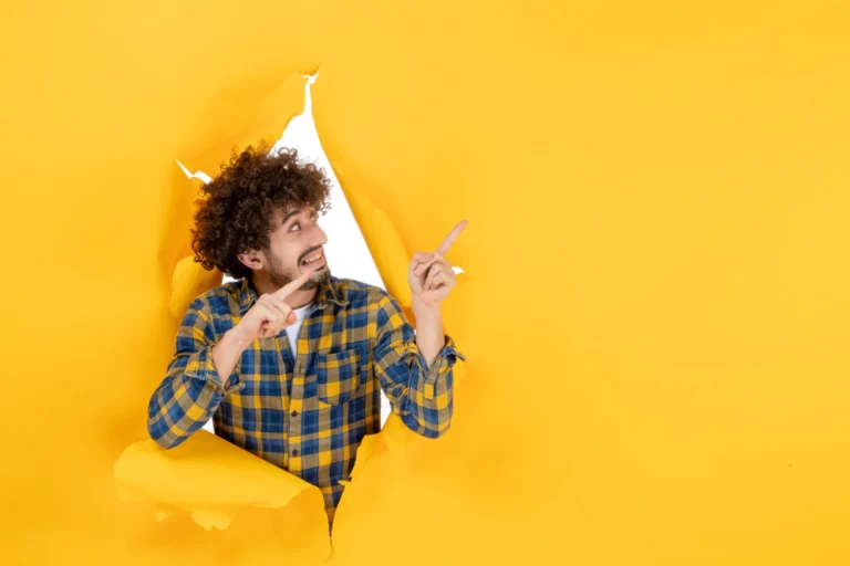 Homem com barba e cabelo cacheado sai de um rasgo na parede amarela e aponta para a lateral, sugerindo uma ideia criativa para Criativos para Anúncios: Como Criar Peças que Geram Interesse e Cliques