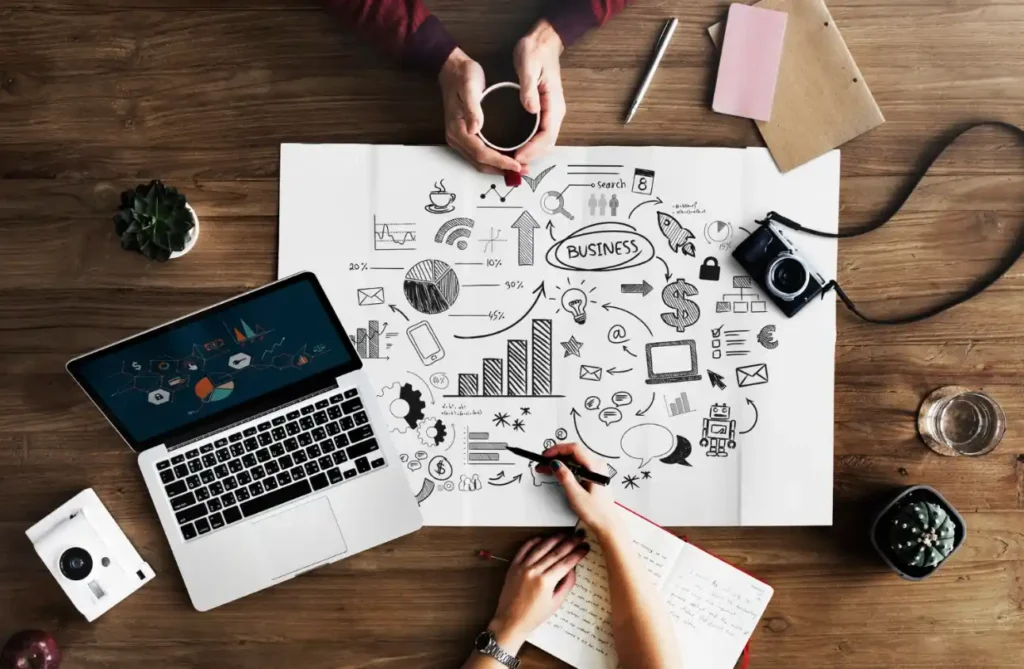 Mesa de trabalho com notebook, celular e câmera ao lado de um desenho de gráficos, setas e ícones de marketing feitos em papel, enquanto mãos analisam um plano. Conceito de Publicidade Digital: Como Criar Campanhas que Chamam a Atenção do Público.